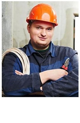 A man wearing an orange hard hat and blue work uniform stands with arms crossed, holding a coil of cable and pliers, representing the pre-eminent electrical service company in Alamance County, NC, in a construction setting.